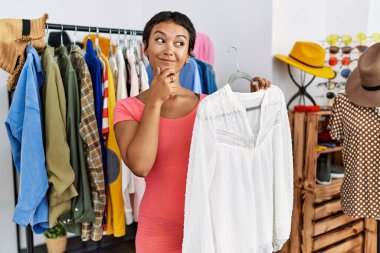 Young hispanic woman with short hair shopping at retail boutique serious face thinking about question with hand on chin, thoughtful about confusing idea 