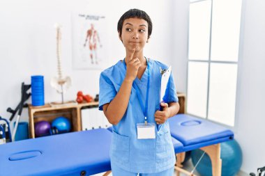 Kısa saçlı, İspanyol bir kadın ağrı kesici kliniğinde çalışıyor. Çene kemiğinde parmak olan şüpheye odaklanmış durumda. 