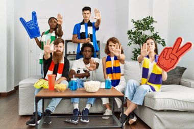 Group of friends supporting football team watching tv al home sitting on the sofa with open hand doing stop sign with serious and confident expression, defense gesture 