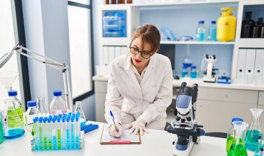 Young caucasian woman wearing scientist uniform writing on clipboard at laboratory
