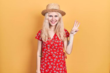 Sarışın, güzel, beyaz bir kadın. Yaz şapkası takıyor. Üç numaralı parmaklarıyla işaret ediyor.. 