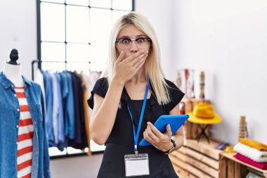 Young blonde woman working as manager at retail boutique shocked covering mouth with hands for mistake. secret concept. 
