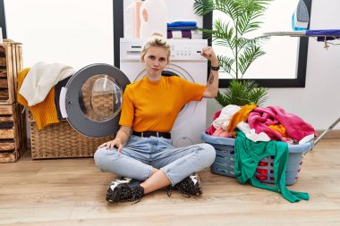 Young blonde woman doing laundry sitting by washing machine strong person showing arm muscle, confident and proud of power 