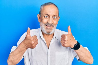 Sakallı, sıradan, beyaz tişörtlü, başarılı bir adam. Elleriyle olumlu bir jest yapıyor. Gülümsüyor ve mutlu. Neşeli ifade ve kazanan jest. 