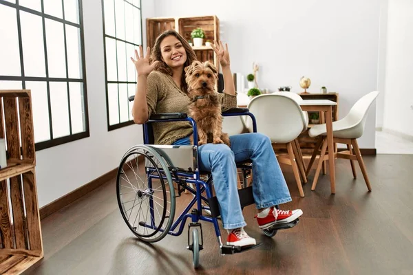 自信と幸せ笑顔ながら家で車椅子に座って指の番号7で指差す若いヒスパニック系の女の子 — ストック写真