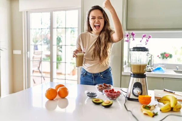 Güzel, genç, esmer bir kadın mutfakta bir bardak smoothie içiyor öfkeyle bağırıyor, öfkeyle bağırıyor ve el kaldırıyor. 