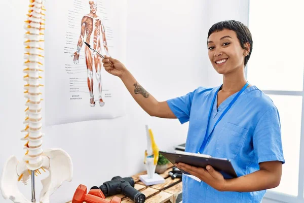 Young hispanic woman wearing physiotherapist uniform pointing to vertebral column holding checklist at physiotherapy clinic