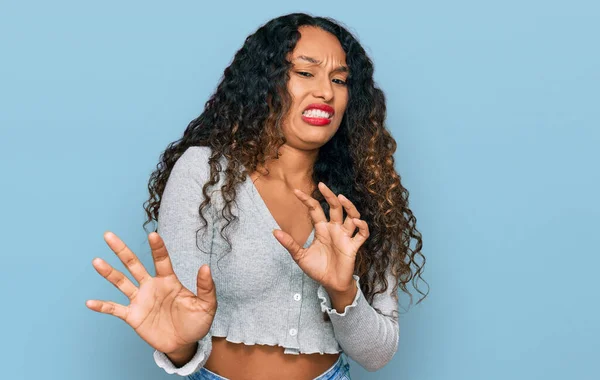 Young hispanic woman with curly hair wearing casual clothes disgusted expression, displeased and fearful doing disgust face because aversion reaction. with hands raised