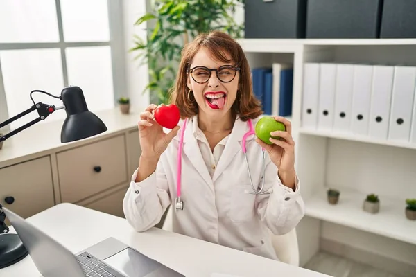 Young Doctor Woman Holding Heart Green Apple Sticking Tongue Out — стоковое фото