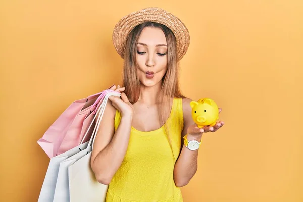 Young blonde girl holding shopping bags and piggy bank making fish face with mouth and squinting eyes, crazy and comical.