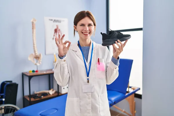 Young Physiotherapist Woman Holding Sneakers Doing Sign Fingers Smiling Friendly —  Fotos de Stock