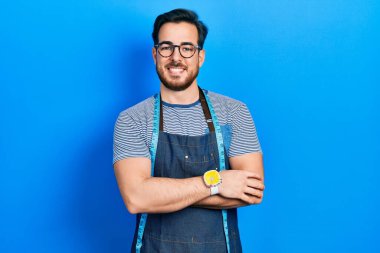 Sakal terzisi olan yakışıklı beyaz adam atölye önlüğü giymiş, kollarını kavuşturup kameraya bakarken gülümsüyor. pozitif kişi. 