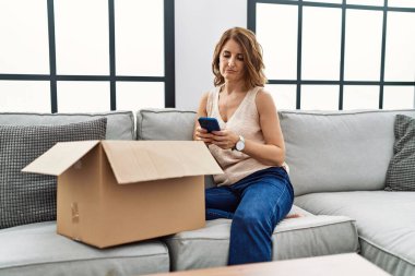 Orta yaşlı bir kadın koltukta oturmuş teslimat paketini telefonla kontrol ediyor. Düşünme tavrı ve ayık yüz ifadesi. Kendine güveniyor gibi görünüyor. 