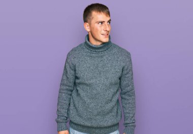 Gündelik kıyafetler giyen genç sarışın adam yüzünde gülümsemeyle yan yana bakıyor, doğal bir ifade. Kendinden emin gülen. 