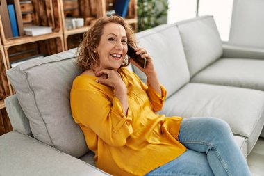 Orta yaşlı beyaz bir kadın akıllı telefonla konuşuyor. Evdeki kanepede oturuyor..