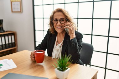 Orta yaşlı beyaz iş kadını ofiste çalışan akıllı telefondan konuşuyor..