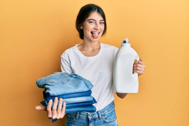 Çamaşır yıkayan beyaz bir kadın elinde deterjan şişesi ve katlanmış kot pantolon ile mutlu bir ifadeyle dilini dışarı çıkarıyor.. 