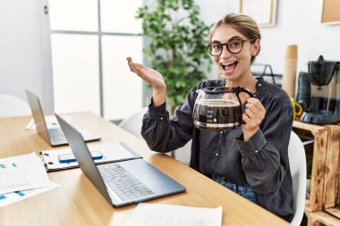 Ofiste çalışan genç sarışın kadın kahve makinesini elinde tutarak başarıyı mutlu bir gülümsemeyle ve kazanan yüz ifadesiyle kutluyor. 