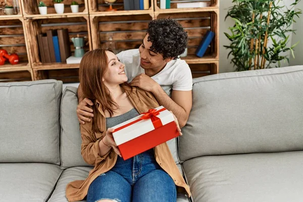 Mutlu gülümseyen genç çift evde yıldönümü hediyesiyle koltukta oturuyor..