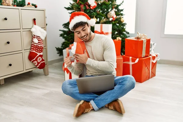 Joven Hombre Hispano Sonriendo Feliz Sosteniendo Regalo Navidad Usando Ordenador — Foto de Stock