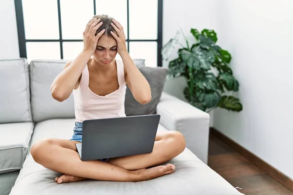 年轻的金发女人在家里用笔记本电脑坐在沙发上 由于疼痛和偏头痛 绝望和紧张 手放在头上 — 图库照片