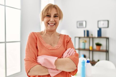 Orta yaşlı bir kadın evinde kollarını kavuşturarak mutlu bir şekilde gülümsüyor..