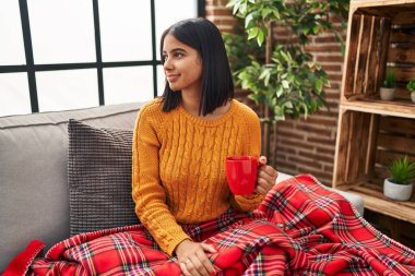 Kanepede oturan Latin kökenli genç bir kadın evde kahve içiyor. Yan tarafa bakıyor, profil pozu veriyor. Doğal bir yüzü ve kendine güvenen bir gülümsemesi var.. 