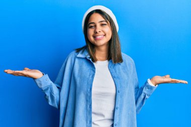 Şirin yün şapkalı genç Latin kadın gülümseyerek avuçlarını açıyor, sunuyor ve reklam karşılaştırması yapıyor ve dengeyi sağlıyor. 