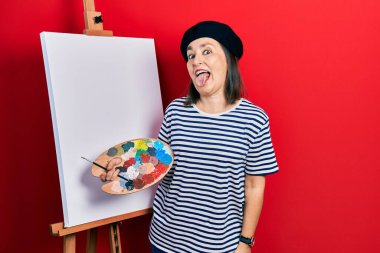 Orta yaşlı İspanyol kadın paletle resim çiziyor, resim sehpası duruyor ve komik bir ifadeyle dilini dışarı çıkarıyor. duygu kavramı. 