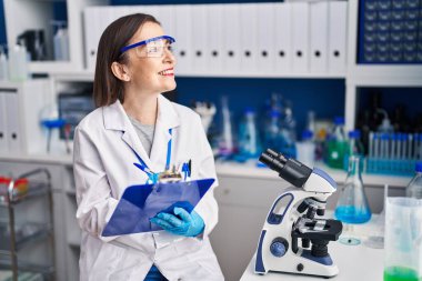 Middle age woman scientist smiling confident writing on clipboard at laboratory