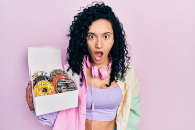 Kıvırcık saçlı genç İspanyol kadın lezzetli renkli donut kutusu tutuyor. Şaşırmak için ağzı açık kalmış, korkmuş ve şaşırmış. İnanmayan yüz. 