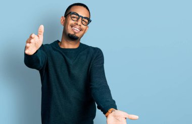 Gündelik giysiler ve gözlük takmış genç Afro-Amerikalı adam kameraya bakıp sarılmak için kollarını açmış gülümsüyor. Mutluluğu kucaklayan neşeli bir ifade.. 
