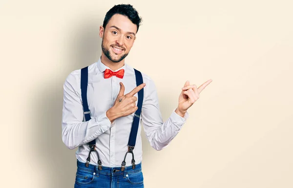 Hispanic man with beard wearing hipster look with bow tie and suspenders smiling and looking at the camera pointing with two hands and fingers to the side.