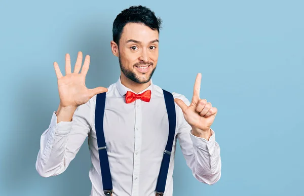 Hispanic man with beard wearing hipster look with bow tie and suspenders showing and pointing up with fingers number seven while smiling confident and happy.