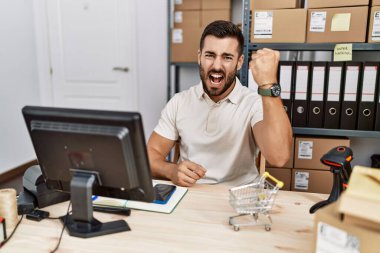 Handsome hispanic man working at small business commerce angry and mad raising fist frustrated and furious while shouting with anger. rage and aggressive concept. 