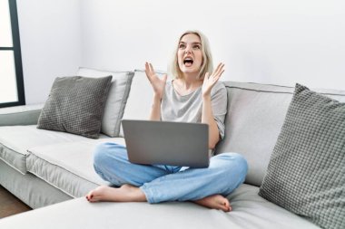 Young caucasian woman using laptop at home sitting on the sofa crazy and mad shouting and yelling with aggressive expression and arms raised. frustration concept. 