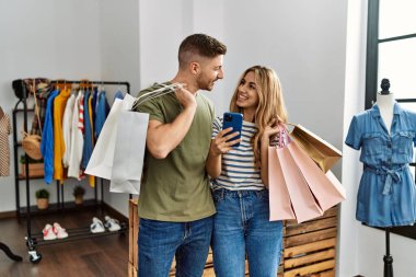 Alışveriş torbaları tutan ve giyim mağazasında akıllı telefon kullanan İspanyol bir çift..