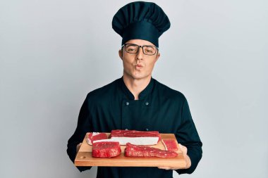 Yakışıklı genç adam, şef üniforması giymiş, elinde çiğ etle kameraya bakıyor ve öpüşüp koklaşıyor. aşk ifadesi. 