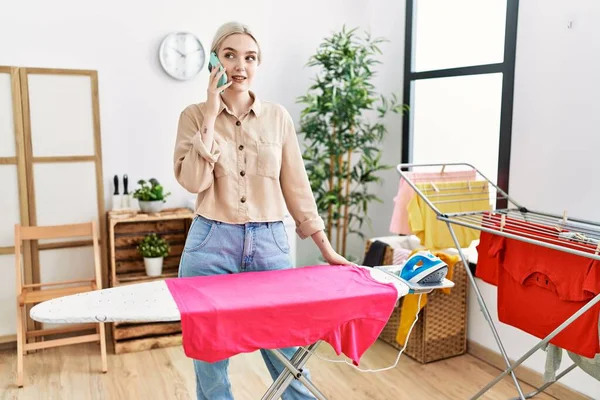 Young Caucasian Woman Talking Smartphone Ironing Clothes Laundry Room — стоковое фото