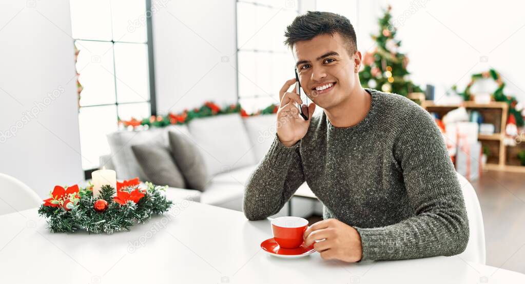Young hispanic man drinking coffee and talking on the smartphone sitting by christmas tree at home