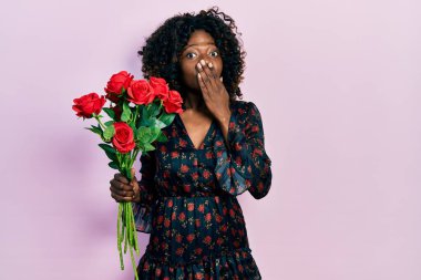 Young african american woman holding bouquet of red roses covering mouth with hand, shocked and afraid for mistake. surprised expression 