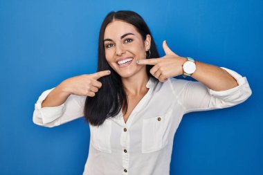 Mavi arka planda dikilen genç İspanyol kadın gülümsüyor ve parmaklarıyla ve ağzıyla işaret ediyor. diş sağlığı kavramı. 