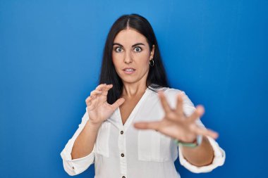 Young hispanic woman standing over blue background afraid and terrified with fear expression stop gesture with hands, shouting in shock. panic concept. 