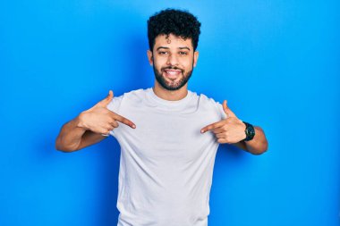 Sakallı, beyaz tişörtlü, kendine güvenen, yüzünde gülümseme olan, kendini gururlu ve mutlu gösteren genç bir Arap.. 