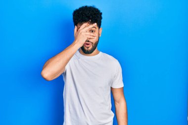 Sakallı genç, beyaz tişörtlü, şokla yüzünü ve gözlerini kapayarak utangaç ifadesiyle parmaklarının arasından bakan Arap bir adam.. 