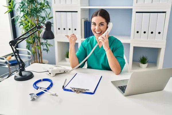 Young Hispanic Doctor Woman Speaking Phone Clinic Pointing Thumb Side — Stock fotografie