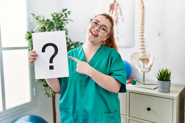 Young Redhead Physiotherapist Woman Holding Question Mark Clinic Smiling Happy — Stock fotografie