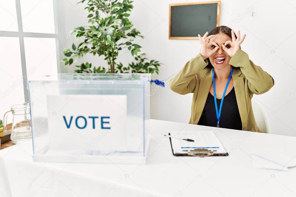 Young brunette woman sitting at election table with voting ballot doing ok gesture like binoculars sticking tongue out, eyes looking through fingers. crazy expression. 