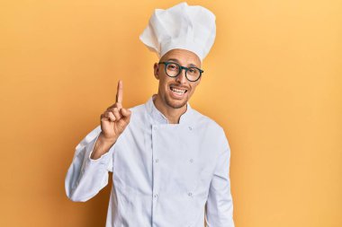 Profesyonel aşçı üniforması giyen kel bir adam bir numarayı gösterip işaret ederken kendinden emin ve mutlu bir şekilde gülümsüyor.. 