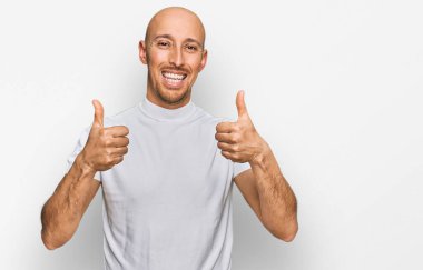 Kel, sakallı, günlük beyaz t-shirt giyen, başarı işareti yapan, el hareketi yapan, gülümseyen ve mutlu bir adam. Neşeli ifade ve kazanan jest. 